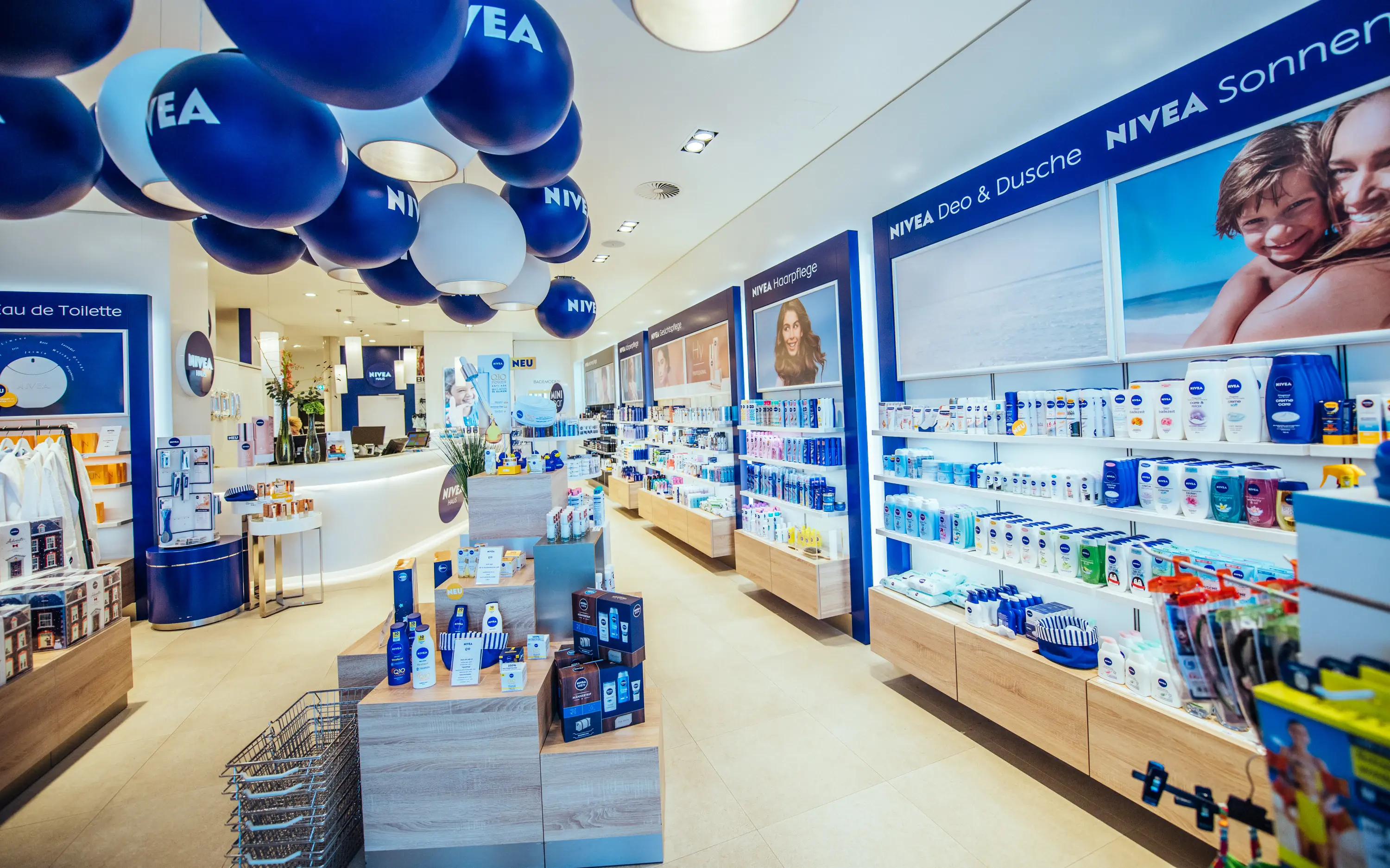 Verkaufsraum NIVEA Haus In einem Verkaufsraum mit hellem Holzboden und weißen Wänden sind diverse NIVEA Produkte in den Regalen zu erkennen. An der Decke hängen blaue und weiße Luftballons mit dem NIVEA Logo bedruckt und im Hintergrund ist ein weißer Verkaufstresen zu erkennen.