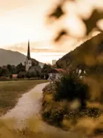 Ein malerischer Pfad führt zu einer historischen Kirche mit spitzem Turm, die in einer idyllischen Landschaft liegt. Die Abendsonne taucht die Szene in warmes Licht, während die umgebenden Hügel und Bäume eine ruhige, friedliche Atmosphäre schaffen.