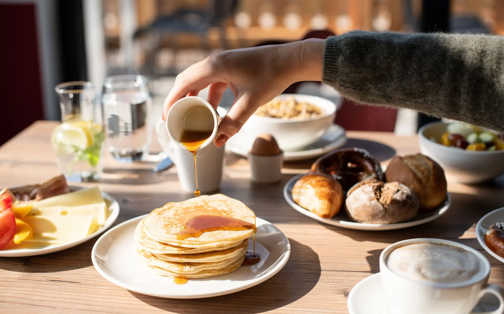 Ein reich gedeckter Frühstückstisch mit Brötchen, Müsli und Pancakes, über die Ahornsirup gegossen wird.