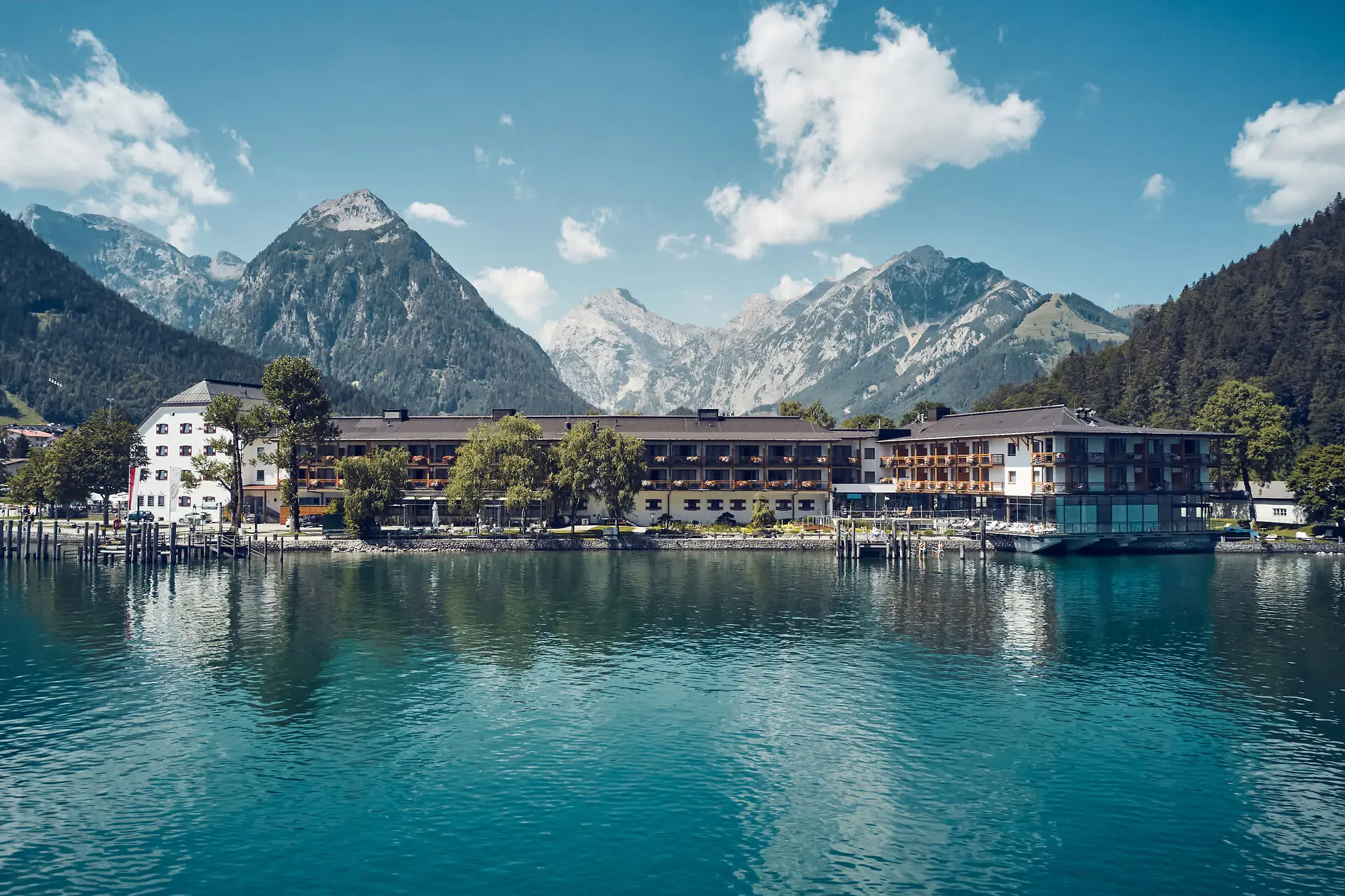 Idyllisches Hotel am Ufer eines klaren Bergsees, umgeben von beeindruckenden Berggipfeln unter einem strahlend blauen Himmel.