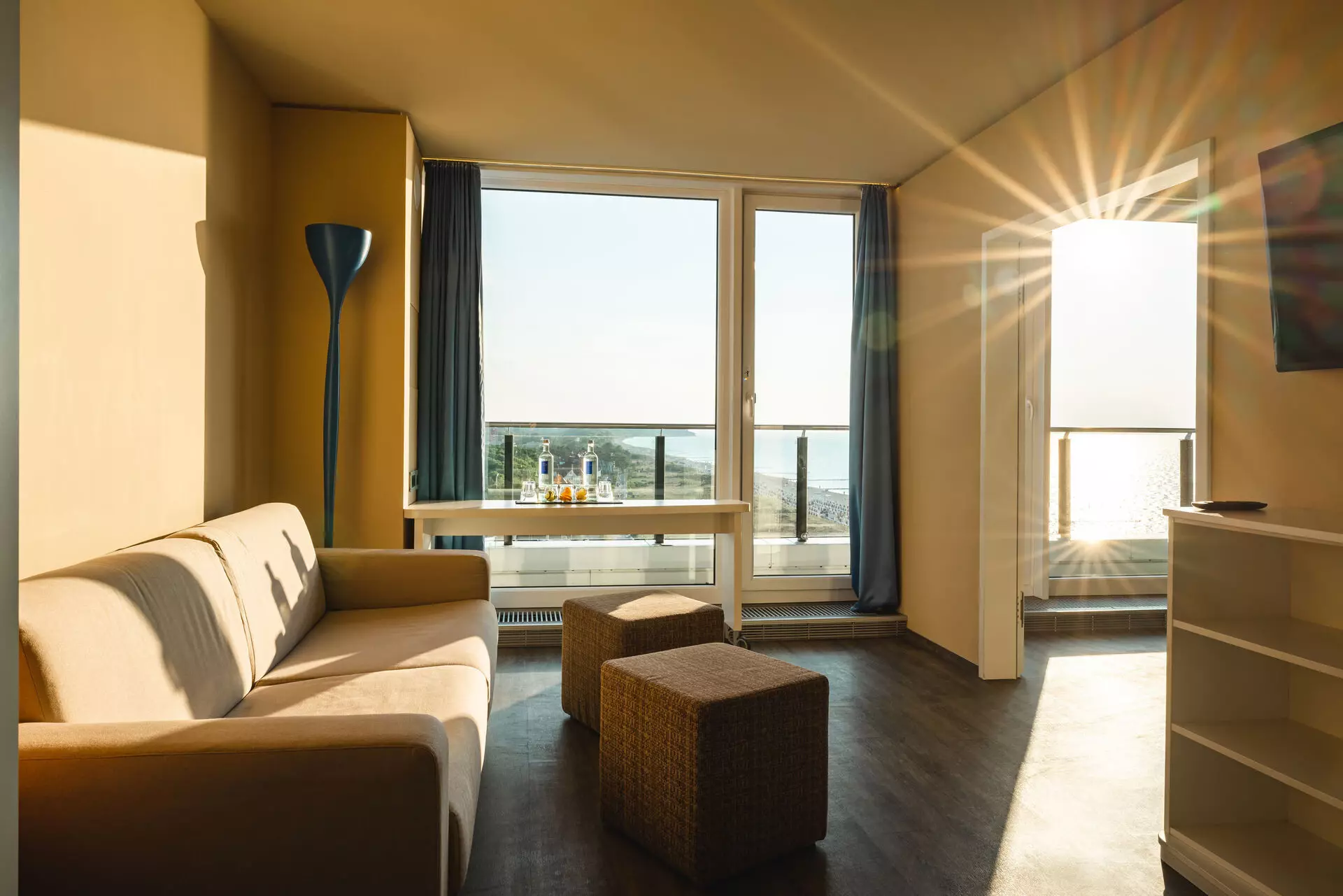 Eine Ausschnitt eines hellen Wohnbereichs in einem Hotelzimmer. Ein helles Sofa steht vor einer weißen Wand an einem Fenster, durch das die Morgensonne in Strahlen in das Zimmer scheint und dieses mit warmem Licht füllt. Gegenüber des Sofas befindet sich eine Kommode und ein Fernseher an der Wand und eine Durchgangstür führt in ein weiteres Zimmer.
