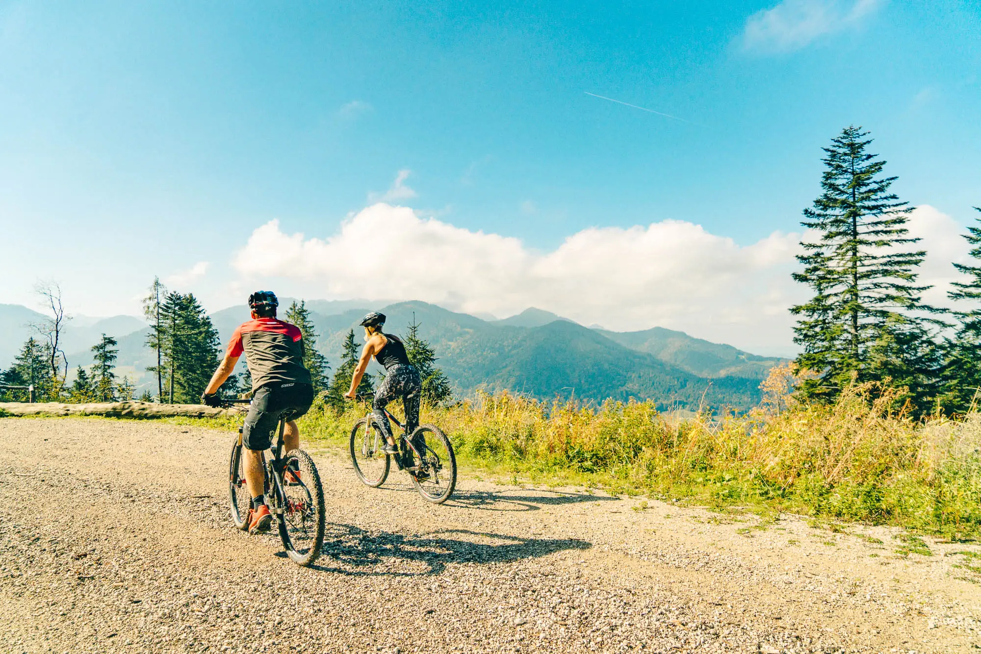 Zwei Personen fahren mit Mountainbikes auf einem Schotterweg inmitten einer alpinen Landschaft, umgeben von Bäumen und Bergen unter einem strahlend blauen Himmel.