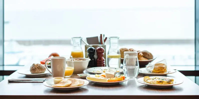 Ein reichlich gedeckter Frühstückstisch an einem Fenster mit Früchten, Pancakes, Brötchen und Getränken. Ein reichlich gedeckter Frühstückstisch an einem Fenster mit Früchten, Pancakes, Brötchen und Getränken.