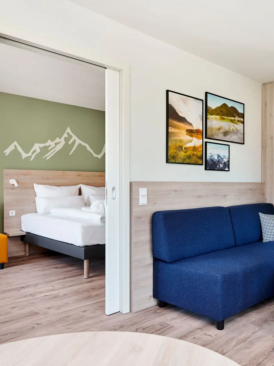 Ein Hotelzimmer mit einem Wohnbereich in dem ein blaues Sofa an einem runden Tisch steht und einem Schlafzimmer im Zimmer dahinter.