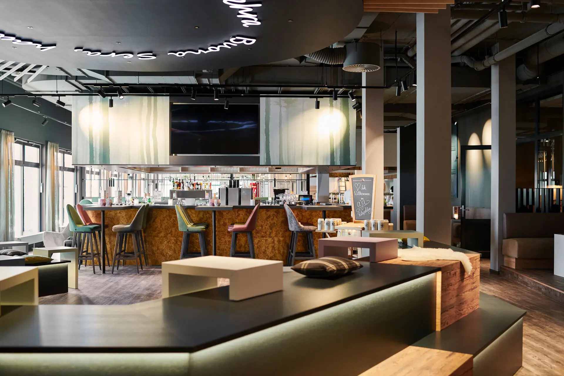 Modernes Bar- und Lounge-Design mit einladendem Ambiente: Stilvolle Barhocker in verschiedenen Farben um eine zentrale Bar, umgeben von warmem Holzdekor und gemütlichen Sitzbereichen.