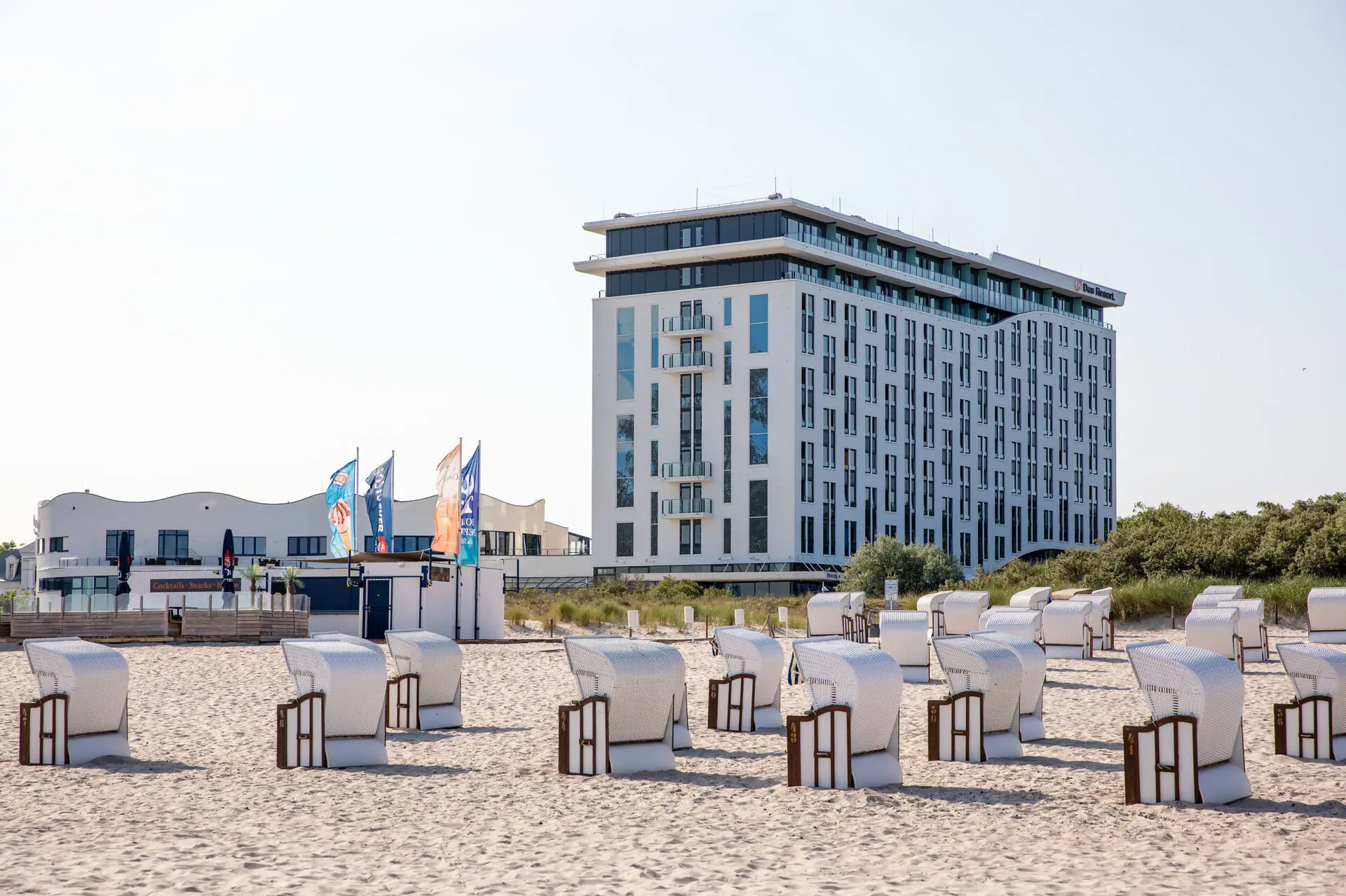 aja Warnemünde Vor einem weißen Gebäude am Strand stehen Strandkörbe aufgereiht und die Sonne scheint.