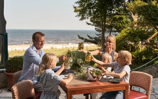 Eine Familie mit drei Kindern sitzt auf einer Terrasse an einem Tisch, auf dem Essen steht und stößt mit Limonaden an. Im Hintergrund ist das Meer und der Strand zu sehen. Eine Familie mit drei Kindern sitzt auf einer Terrasse an einem Tisch, auf dem Essen steht und stößt mit Limonaden an. Im Hintergrund ist das Meer und der Strand zu sehen.
