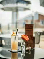 Auf einem grauen runden Tisch auf einer Terrasse steht ein Cocktail in einem hohen Glas mit Strohhalm neben einer kleinen Vase mit einer orangefarbenen Blume und braunen Getränkekarten.