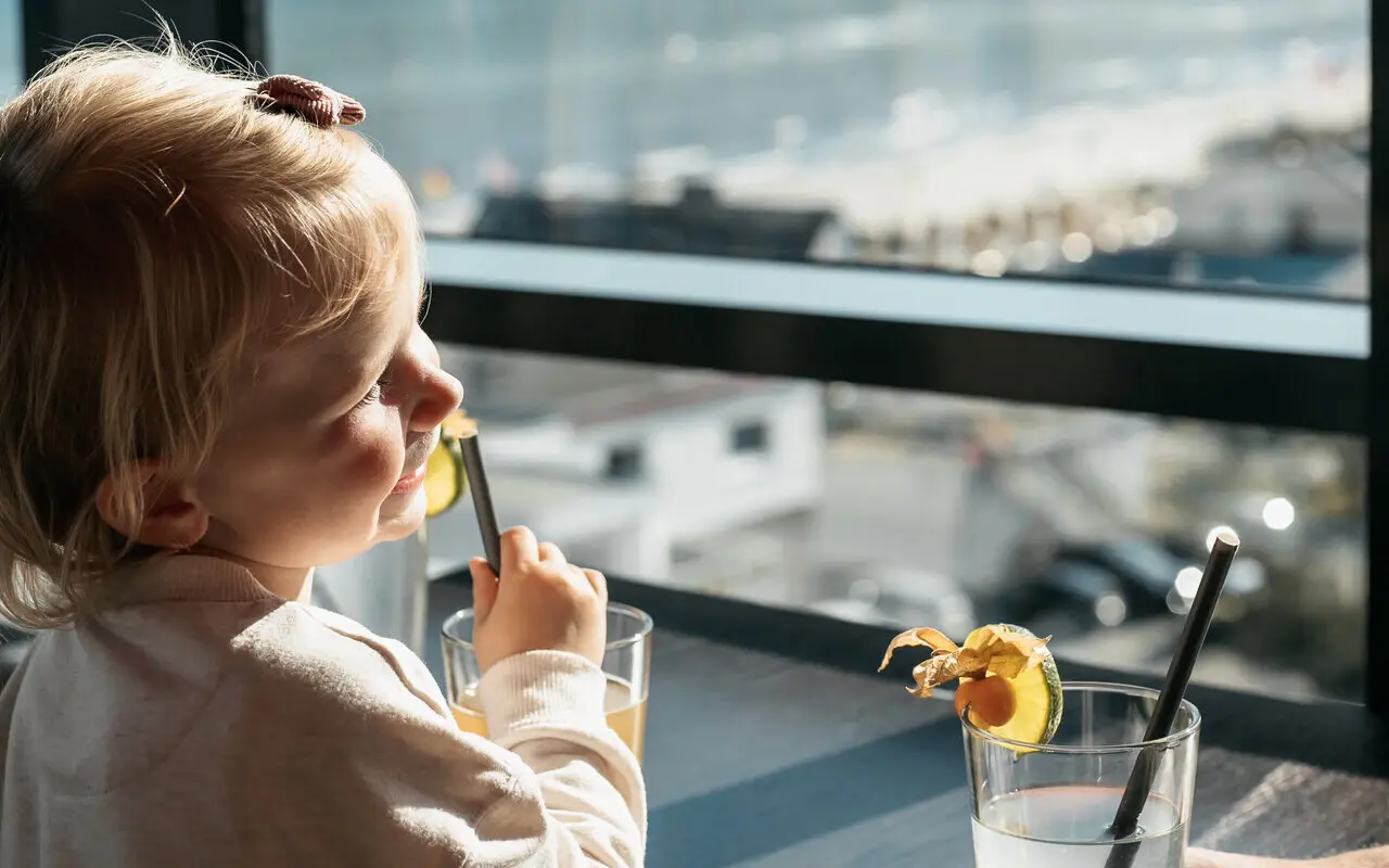 Kind mit Getränk Ein kleines Mädchen sitzt an einer Fensterfront in der Sonne und trinkt aus einem Glas mit einem Strohhalm.