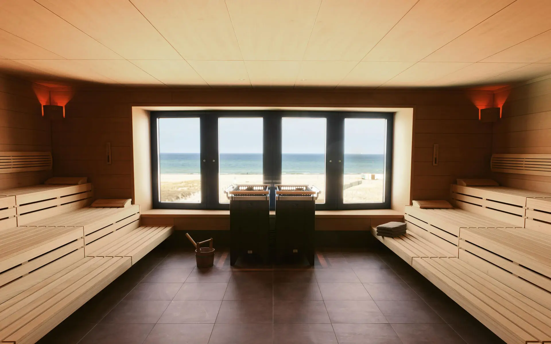 Eine geräumige Holzsauna mit breiter Fensterfront die auf den Strand und die angrenzende Ostsee blickt. Vor dem Fenster steht ein großer Saunaofen und ein Holzeimer mit Kelle daneben.