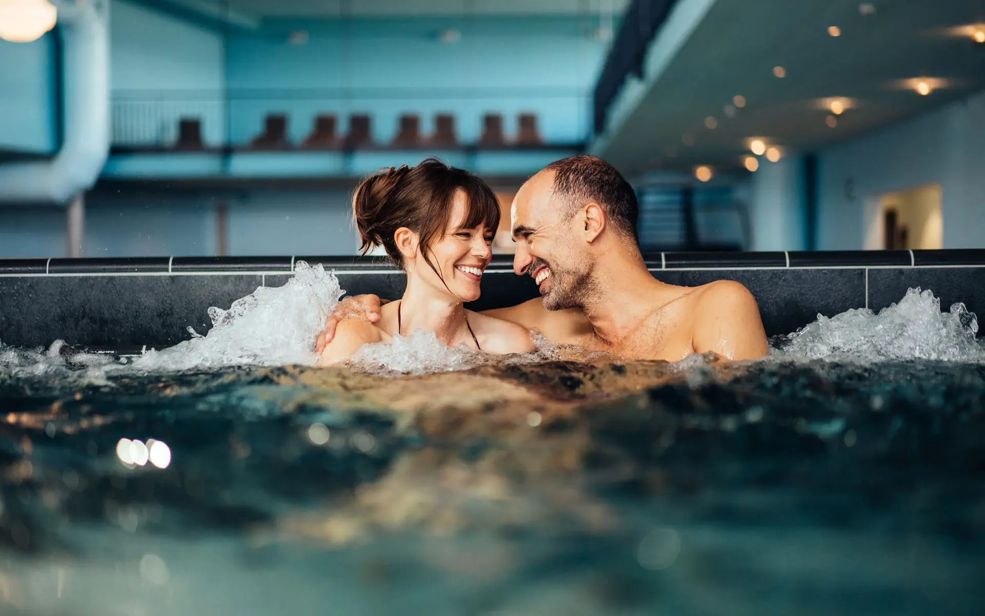 Whirlpool aja Ruhpolding Ein Mann und eine Frau sitzen in einem sprudelnden Schwimmbecken. Der Mann hat den Arm um die Schulter der Frau gelegt und sie lächeln sich an, die Blicke einander zugewandt. Im Hintergrund ist abgedunkelt und leicht verschwommen einige Liegen im oberen Bereich zu erkennen.
