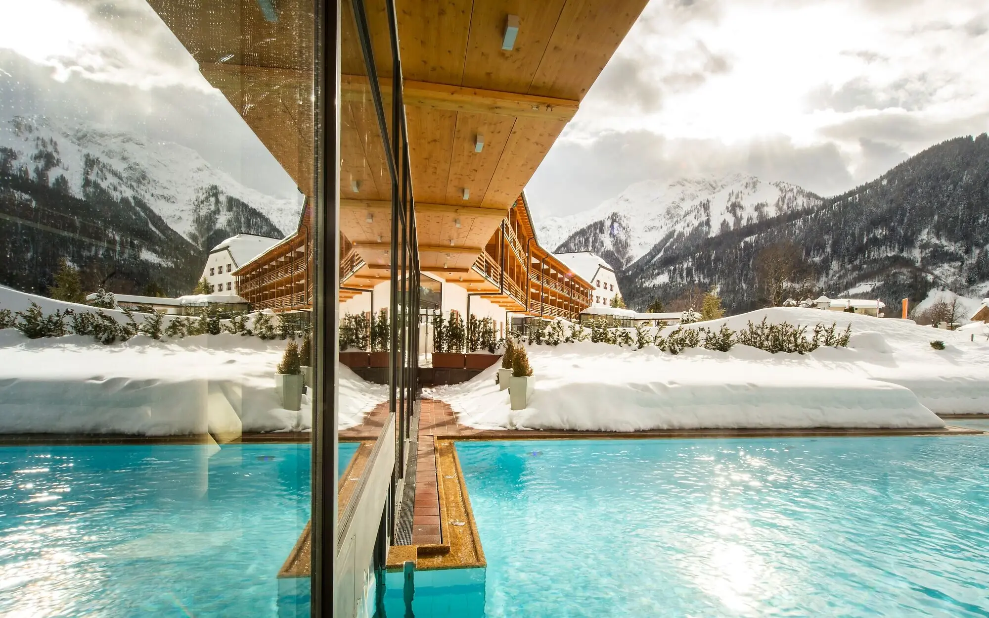 Außenpool aja Achensee Ein beheizter Außenpool in einer verschneiten Winterlandschaft, flankiert von einer modernen Glasfassade und einem Holzvordach. Im Hintergrund ragen majestätische Berge empor