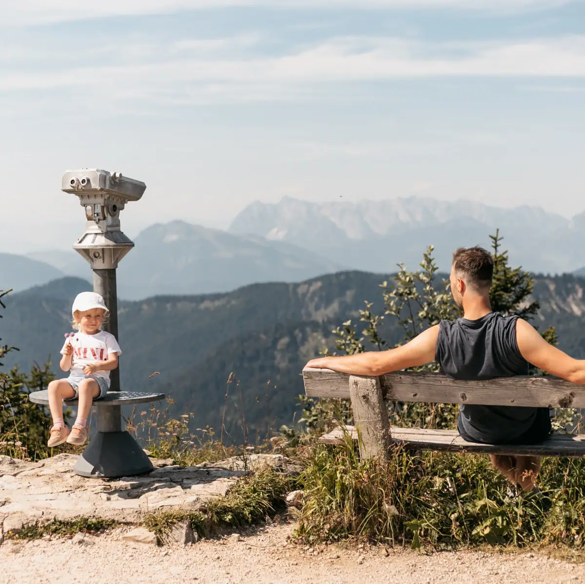 Vater und Tochter genießen die Aussicht auf die Alpen vom Erlebnisberg Rosnerköpfl in Werfenweng. Das Mädchen sitzt auf einem Aussichtsturm, der Mann entspannt auf einer Holzbank mit Blick auf das Bergpanorama.