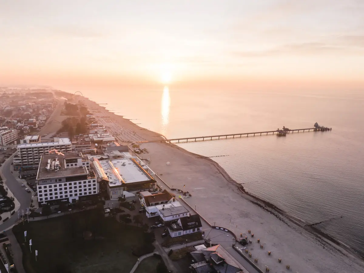 Grömitz Ein Strand mit einem Pier und Gebäuden bei Sonnenuntergang.