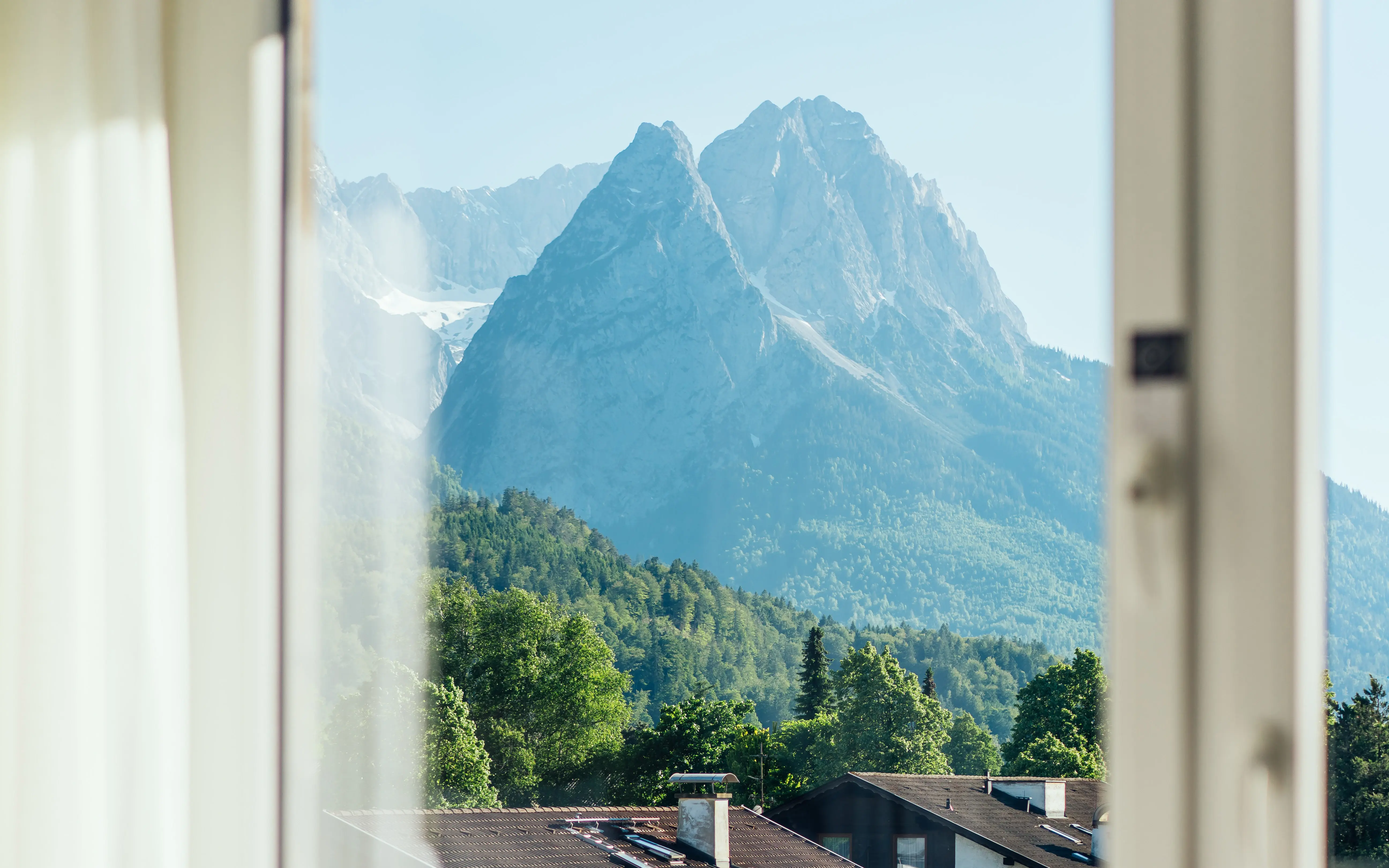 Durch ein großes Fenster hindurch ist eine grün bewachsene Berglandschaft hinter Häusern in der Sonne zu sehen.