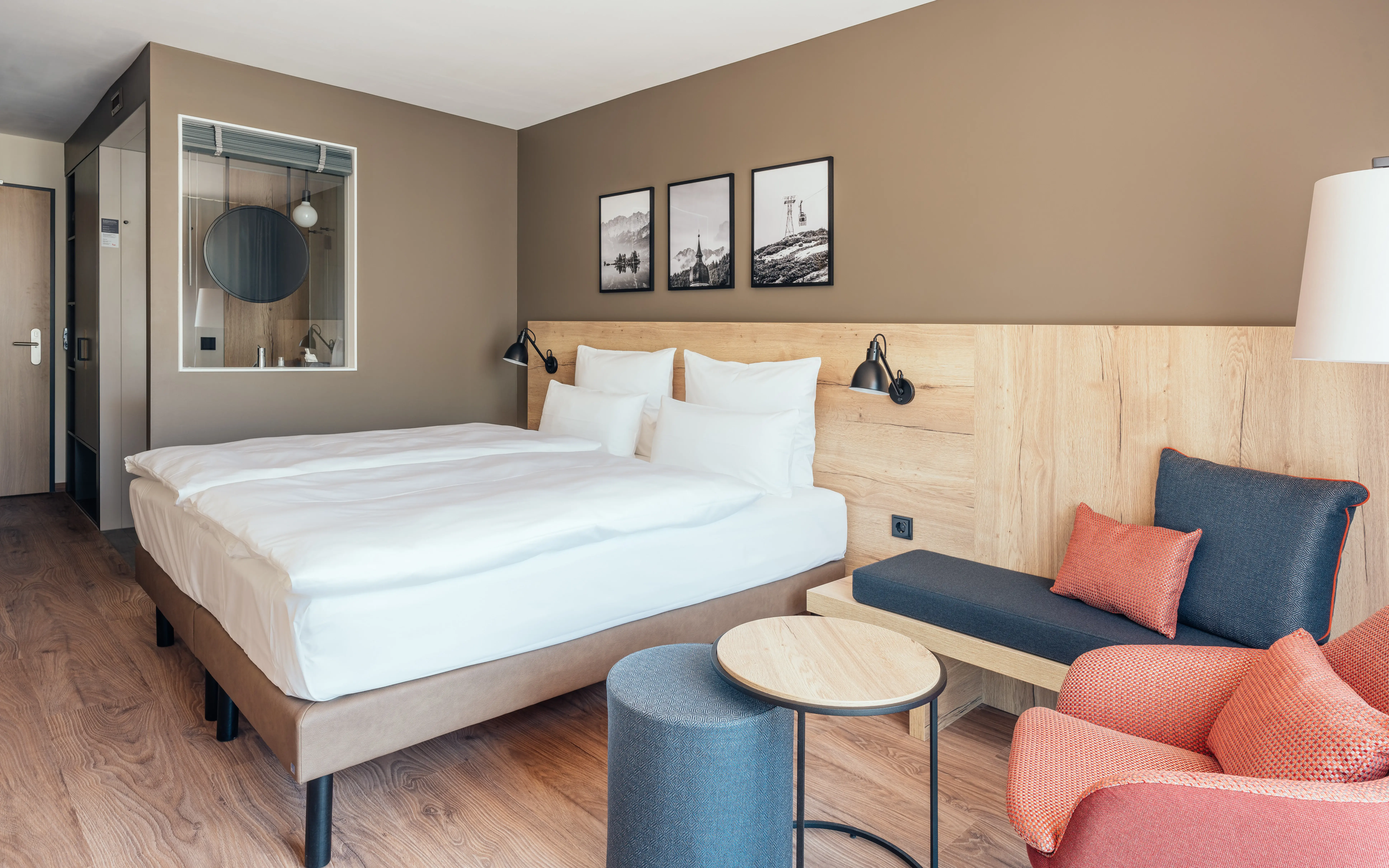 Modernes Hotelzimmer mit gemütlichem Doppelbett, einer Sitzecke und warmen Holzakzenten. An der Wand über dem Bett hängen schwarz-weiße Landschaftsfotografien.Ein roter Sessel und passende Kissen auf der Sitzbank sorgen für Farbakzente und durch ein Fenster ist das badezimmer zu sehen.