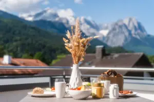 Frühstückstisch auf einer Terrasse mit Bergblick. Verschiedene Speisen wie Brötchen, Brezeln, Obst und Marmelade sind auf dem Tisch arrangiert, dazu eine Vase mit Trockenblumen. Im Hintergrund ragen Berge in den blauen Himmel, die eine idyllische, entspannte Atmosphäre schaffen.