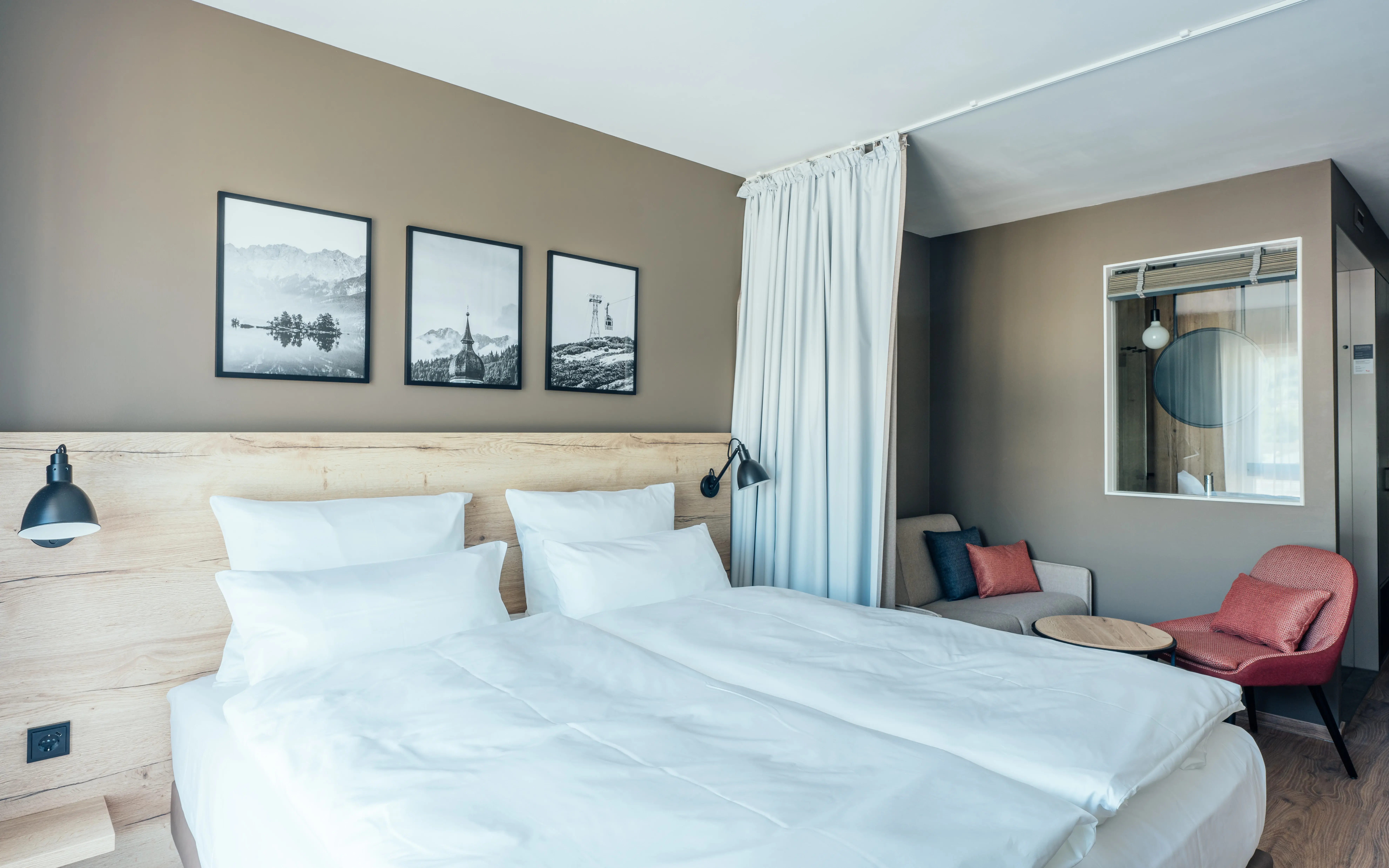 Stilvolles Hotelzimmer mit großem Doppelbett, gemütlicher Sitzecke und natürlichen Holzakzenten. Über dem Bett hängen drei schwarz-weiße Landschaftsbilder.Ein Vorhang bietet zusätzlichen Sichtschutz. Im Hintergrund sind ein roter Sessel, ein Sofa mit farblich abgestimmten Kissen und ein Fenster zum Badezimmer zu sehen.