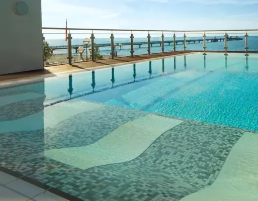 Ausblick über das ruhige Wasser des Außenpools der Grömitzer Welle auf die Ostsee im Hintergrund, welche durch eine gläserne Umzäunung vom Außenbereich des Schwimmbads abgetrennt wird.