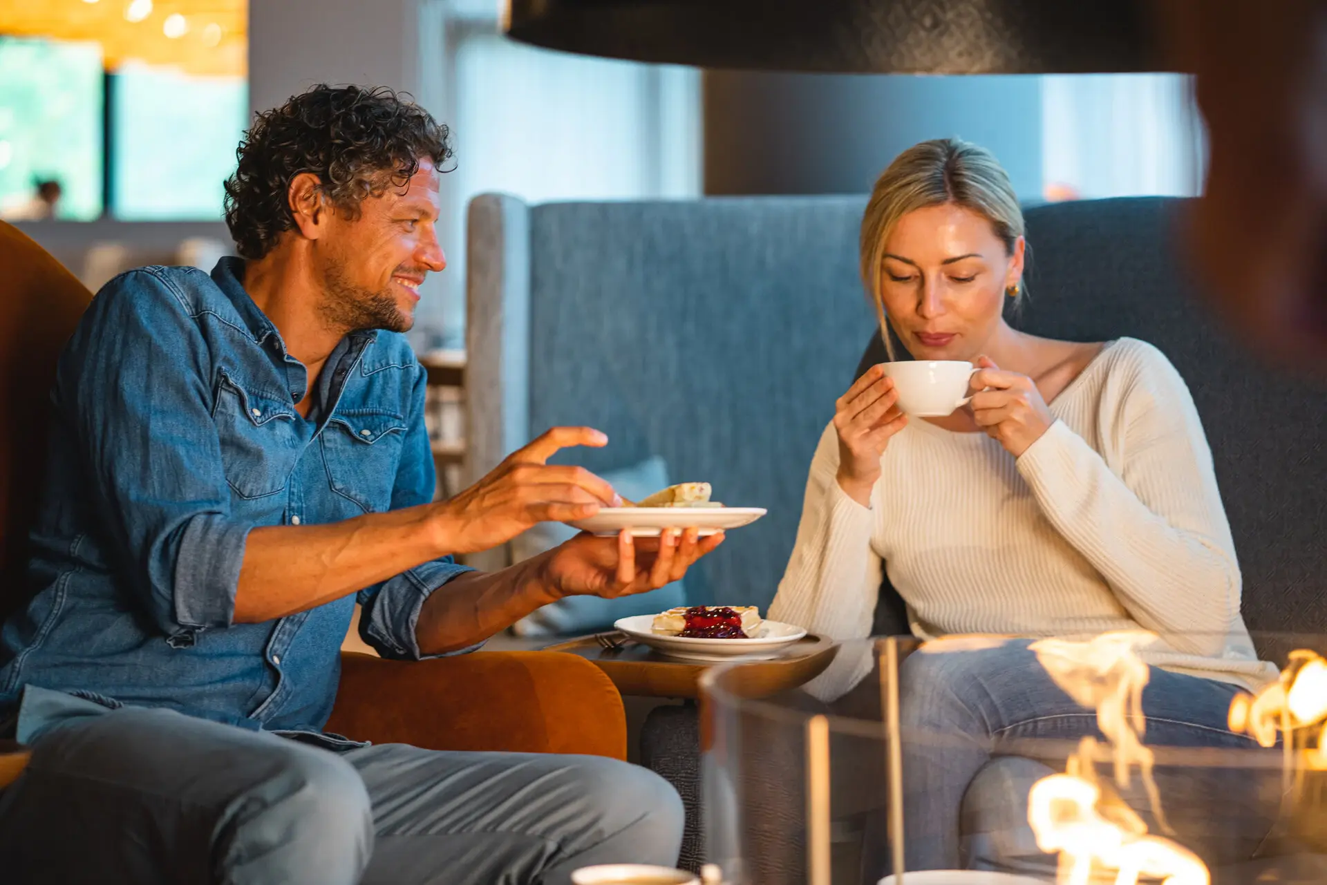 Essen im aja Warnemünde Ein Paar sitzt entspannt in einer gemütlichen Lounge. Der Mann bietet der Frau ein Dessert an, während sie eine Tasse Tee oder Kaffee genießt.