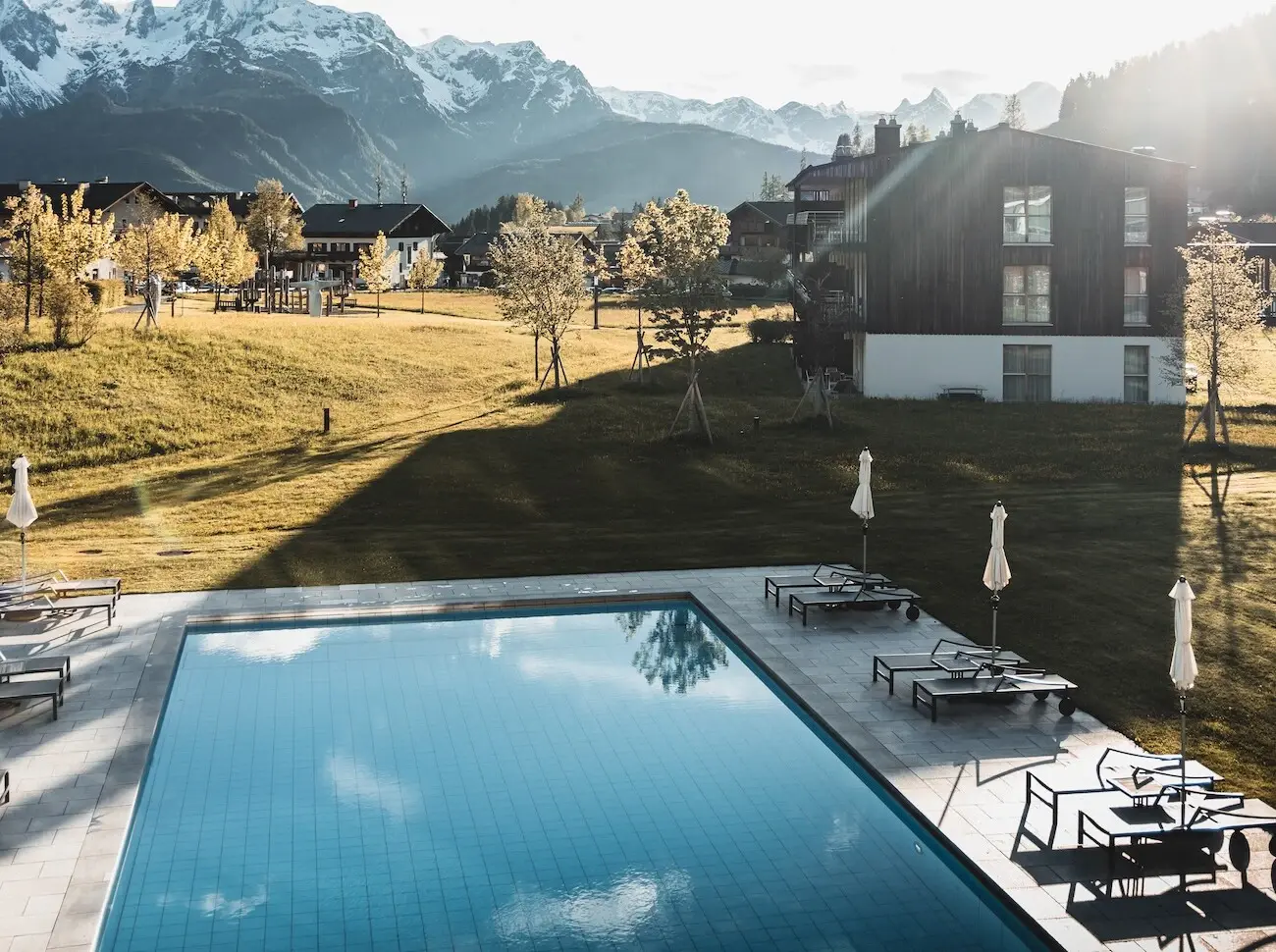 aja Werfenweng Swimmingpool mit Stühlen und Sonnenschirmen vor dem Bergresort Werfenweng mit Bergen im Hintergrund.