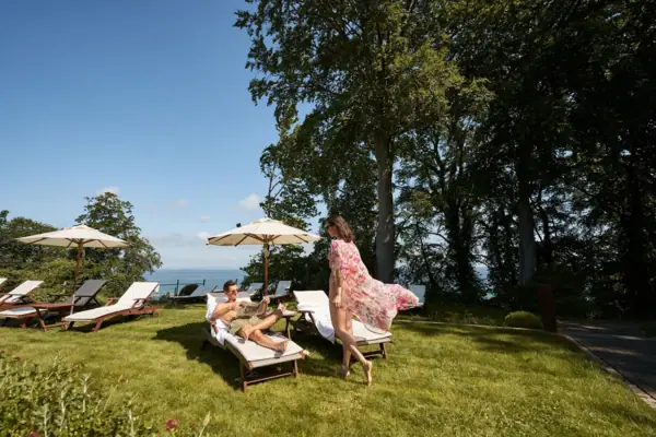 SPA Liegewiese Paar entspannt auf Sonnenliegen im Garten eines Hotels mit Ostseeblick; die Frau trägt ein luftiges Sommerkleid, der Mann prostet mit einem Getränk – Sonnenschirme und grüne Bäume im Hintergrund.