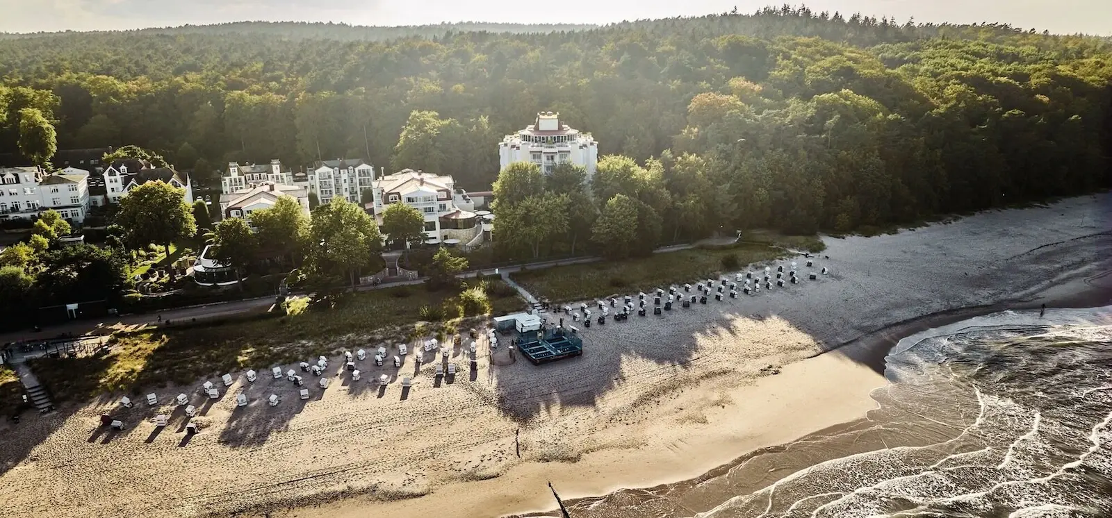 aja Bansin Eine Küstenlandschaft von oben mit vielen Strandkörben am Strand und einigen Häusern zwischen vielen grünen Bäumen.