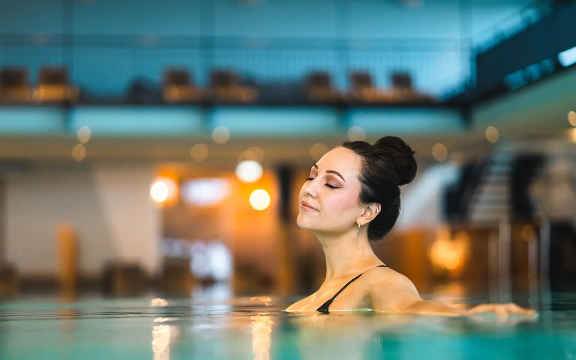 Innenpool aja Travemünde Eine Frau entspannt sich im warmen Wasser eines stilvoll beleuchteten Innenpools. Sie lehnt sich zurück, genießt den Moment mit geschlossenen Augen, während die harmonische Atmosphäre des Wellnessbereichs Ruhe und Gelassenheit ausstrahlt.