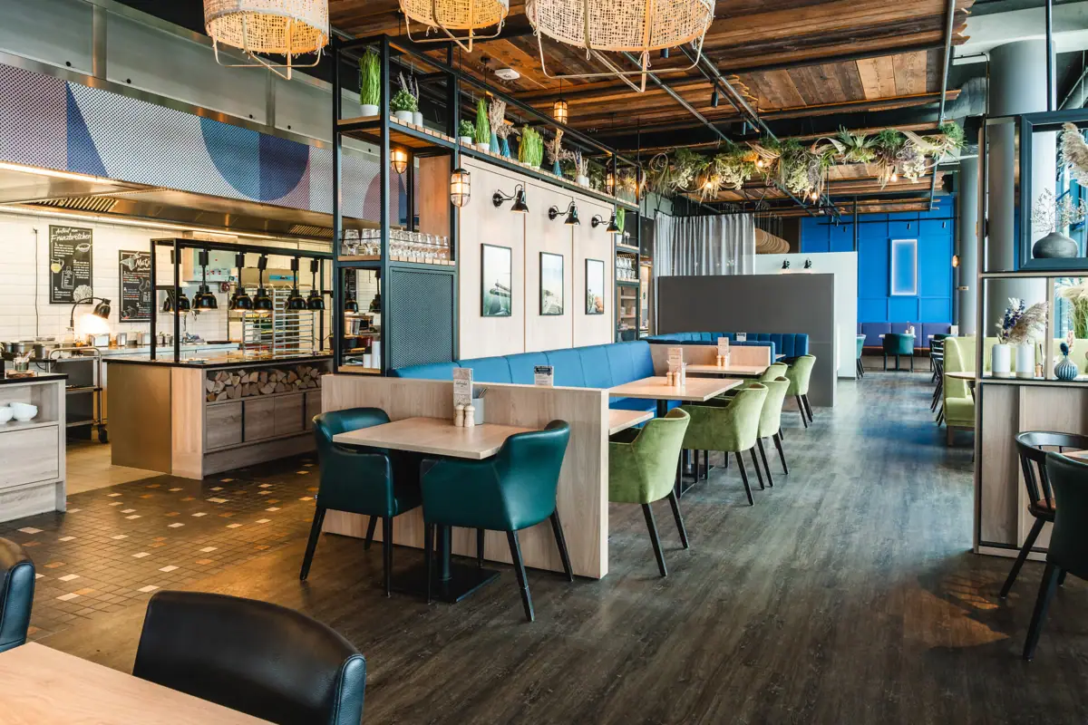 Restaurant im aja Warnemünde Ein Restaurant mit Tischen und Stühlen. Im Hintergrund ist das offene Buffet mit Lampen zu sehen.