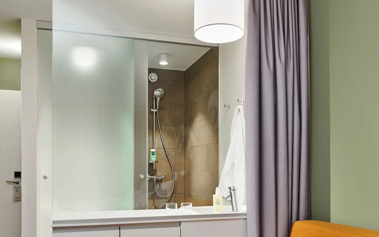 Ein Badezimmer mit Glaswand, hinter der sich die Dusche und das Waschbecken befindet. Ein hellgrauer Vorhang, der als Sichtschutz dient, ist zurückgezogen.