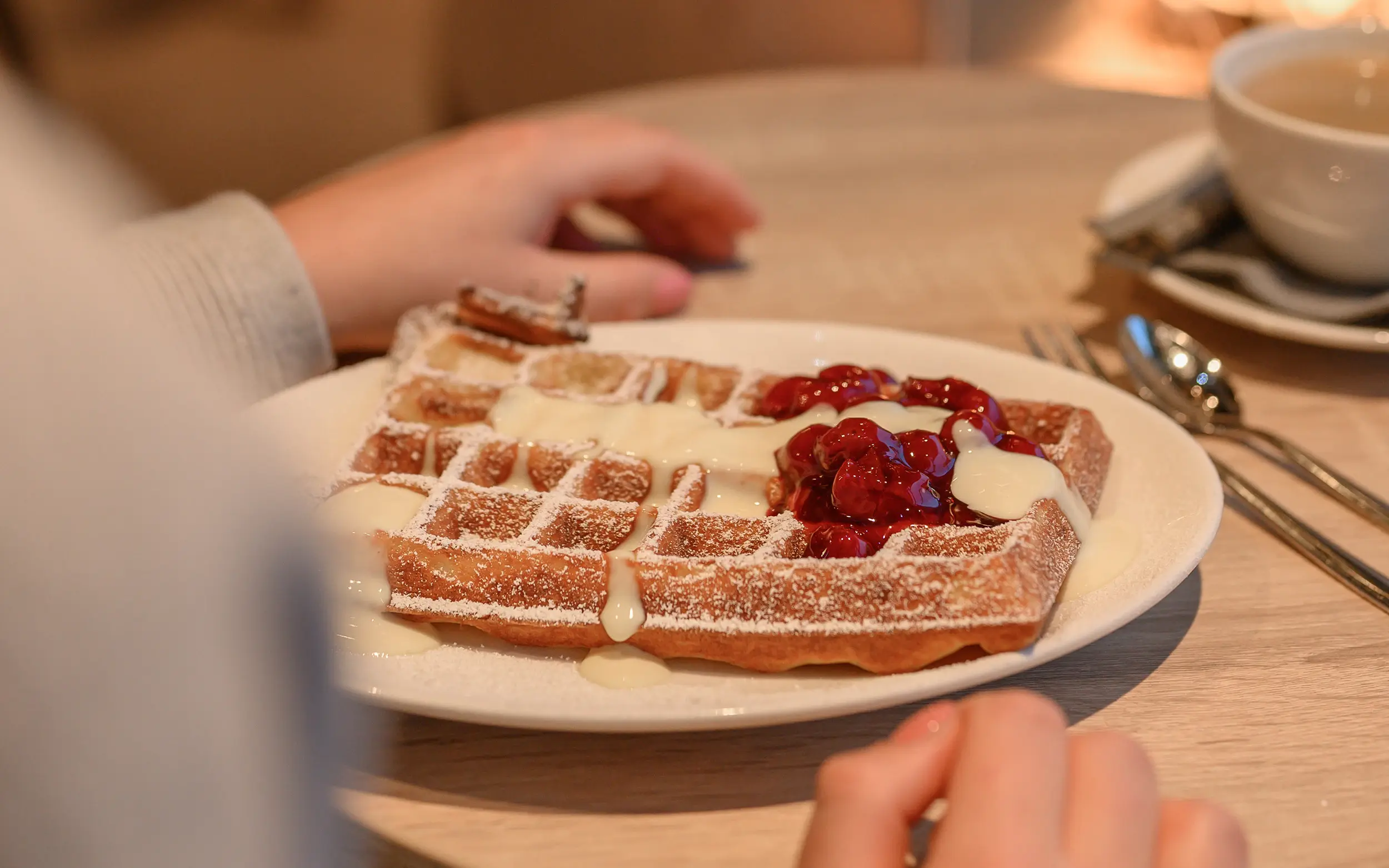 Auf einem Tisch steht vor einer Person ein Waffeletller mit Vanillesauce und Beeren.