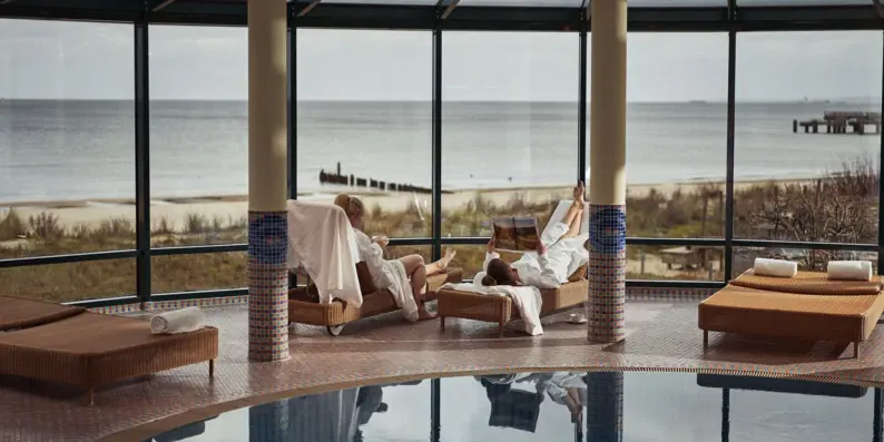 Zwei Personen entspannen sich auf Liegen in einem lichtdurchfluteten Wellnessbereich mit Blick auf den Strand und die Ostsee. Zwei Personen entspannen sich auf Liegen in einem lichtdurchfluteten Wellnessbereich mit Blick auf den Strand und die Ostsee.