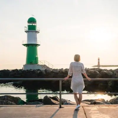 Mole Warnemünde Eine Frau steht auf einem Steg neben einem grün-weißen Leuchtturm.