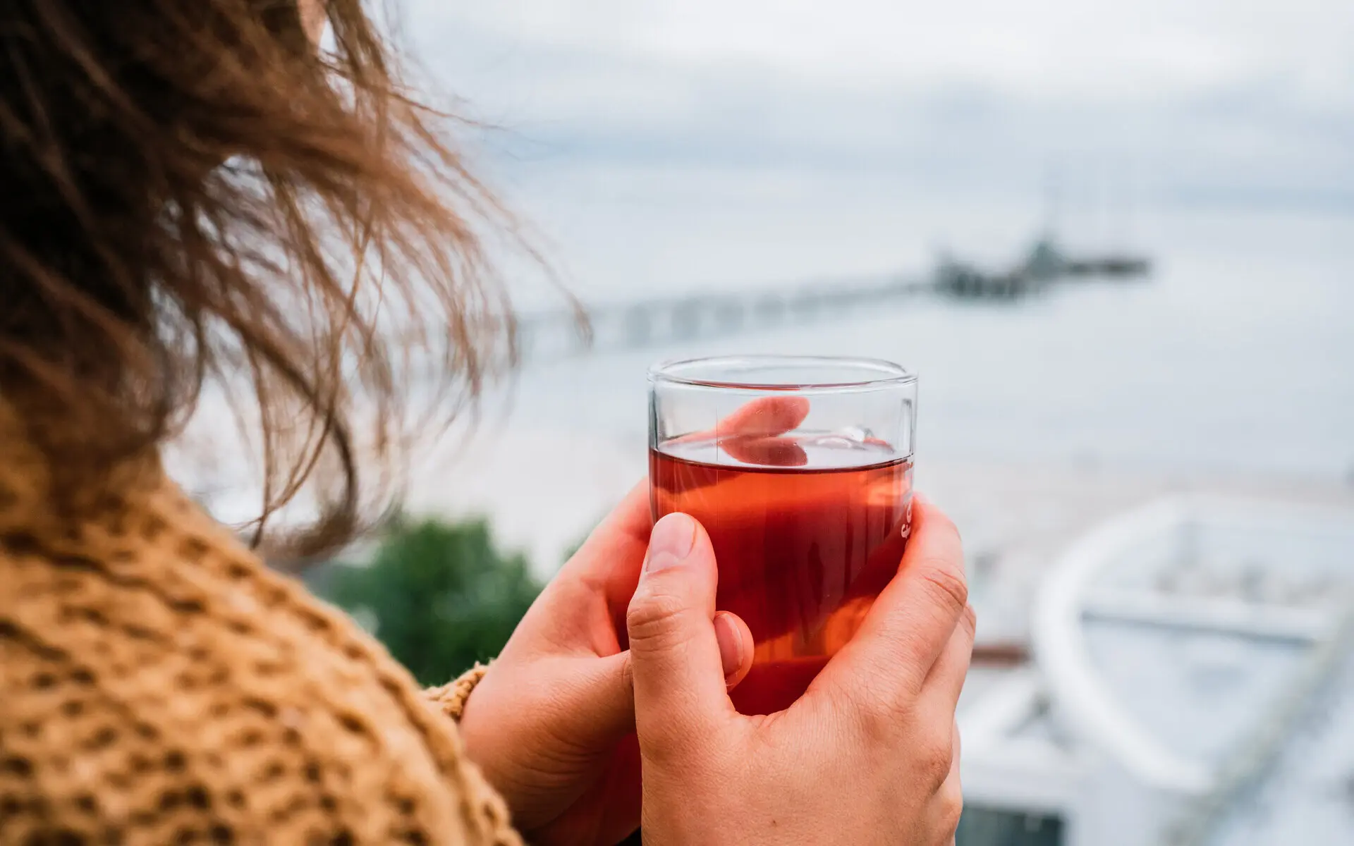 Tee mit Ausblick Ein Frau in Strickpullover steht auf einer windigen Terrasse und hält ein Glas mit einem roten Tee in ihren Händen, während sie auf das Meer in der Ferne schaut.