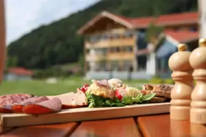Draußen liegt auf einem Holztisch eine Platte mit Aufschnitt. Daneben stehen zwei hölzernen Salz- und Pfefferstreuer und im Hintergrund sind Berge und ein davor stehendes Gebäude.