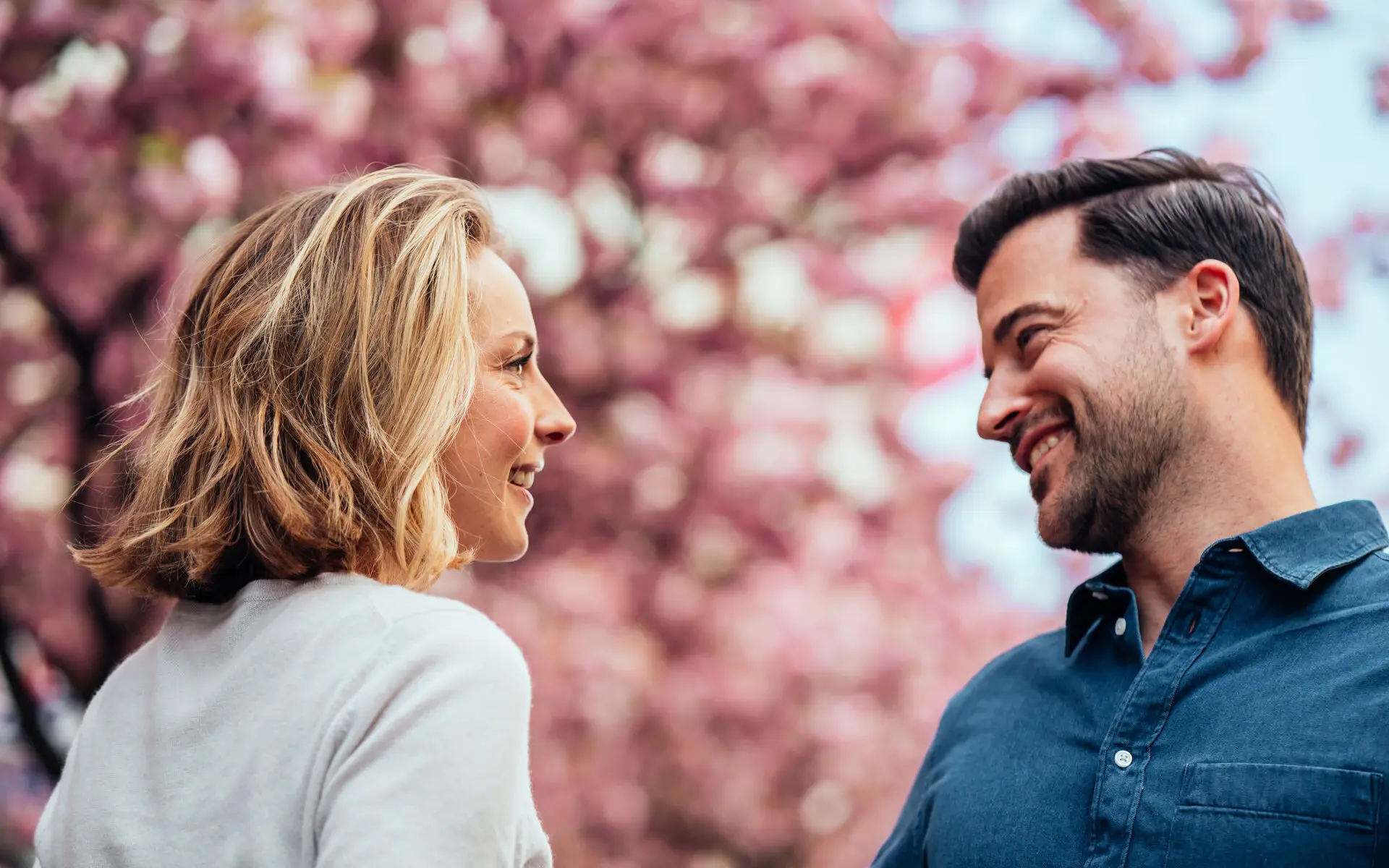Ein Mann und eine Frau stehen sich vor einem rosafarben blühenden Baum gegenüber und lächeln isch an.