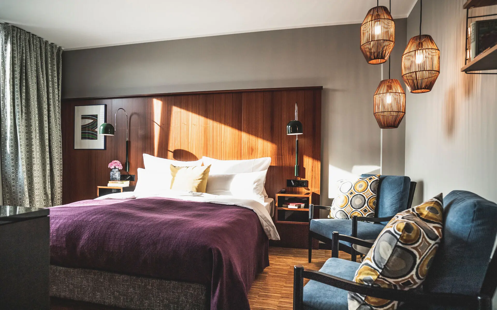 HENRI Düsseldorf Stilvolles Zimmer im HENRI Düsseldorf mit einem gemütlichen Bett, einer violetten Tagesdecke, eleganten Holzelementen, modernen Lampen, blauen Sesseln mit dekorativen Kissen und warmem Tageslicht, das den Raum durchflutet.