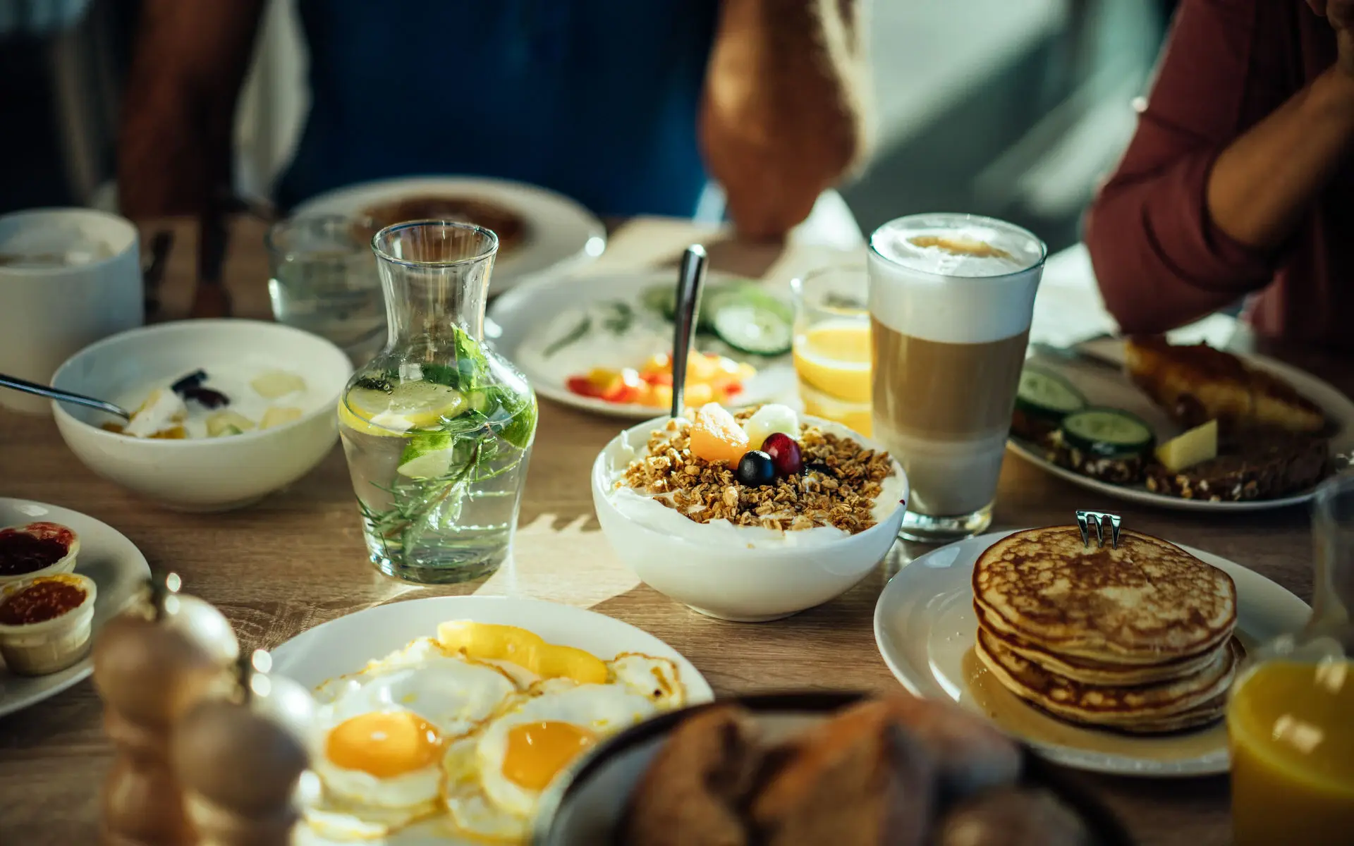 Obstsalat Ein reichlich gedeckter Tisch mit Spiegeleiern, Müsli, Pancakes, Brötchen und Aufschnitt sowie Getränken.