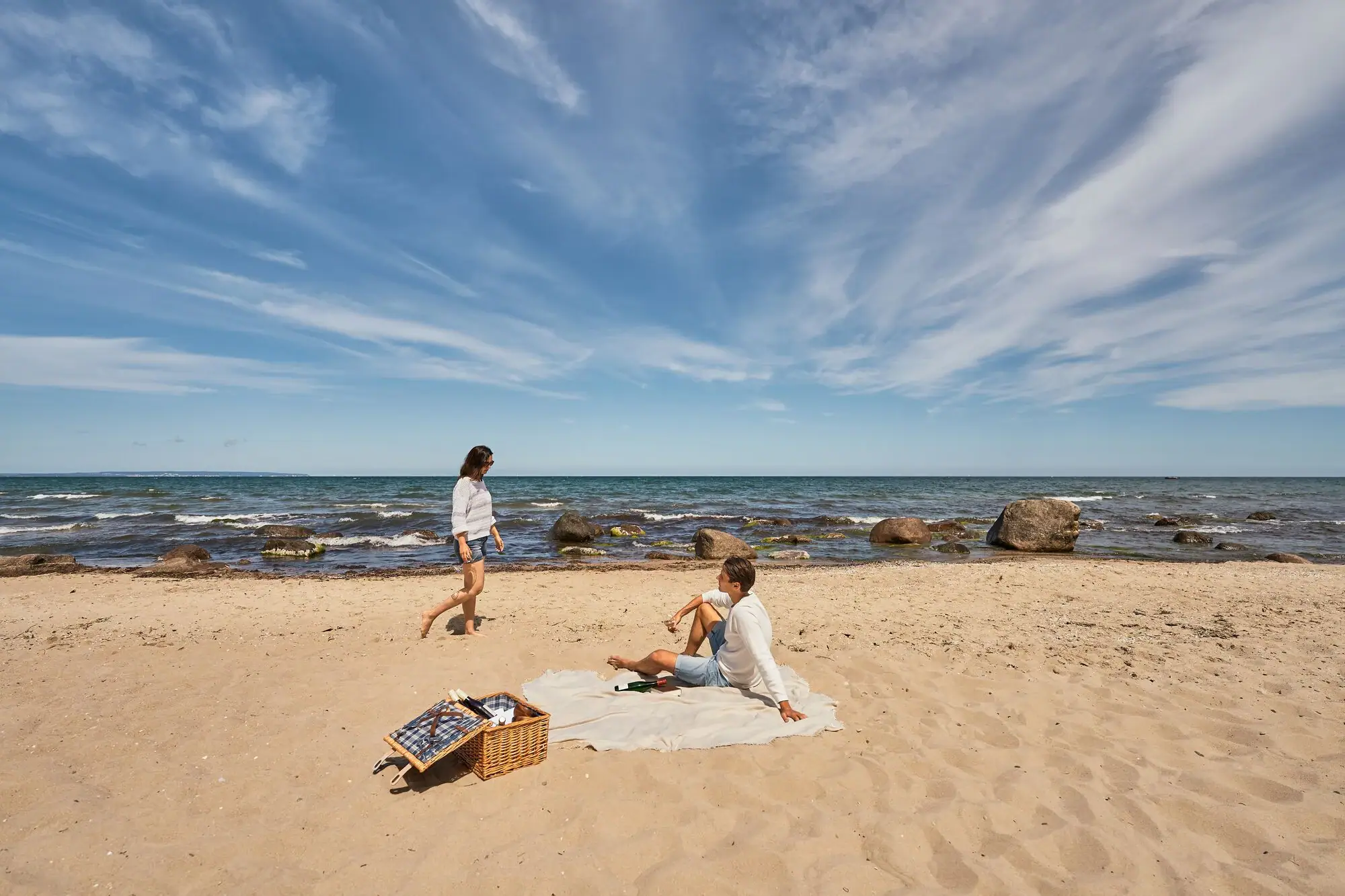 Strand Göhren Paar beim Picknick am naturbelassenen Ostseestrand; der Mann sitzt auf einer Decke im Sand, die Frau spaziert barfuß am Wasser entlang, im Vordergrund ein Picknickkorb, im Hintergrund das Meer mit Felsen und weiter Horizont.