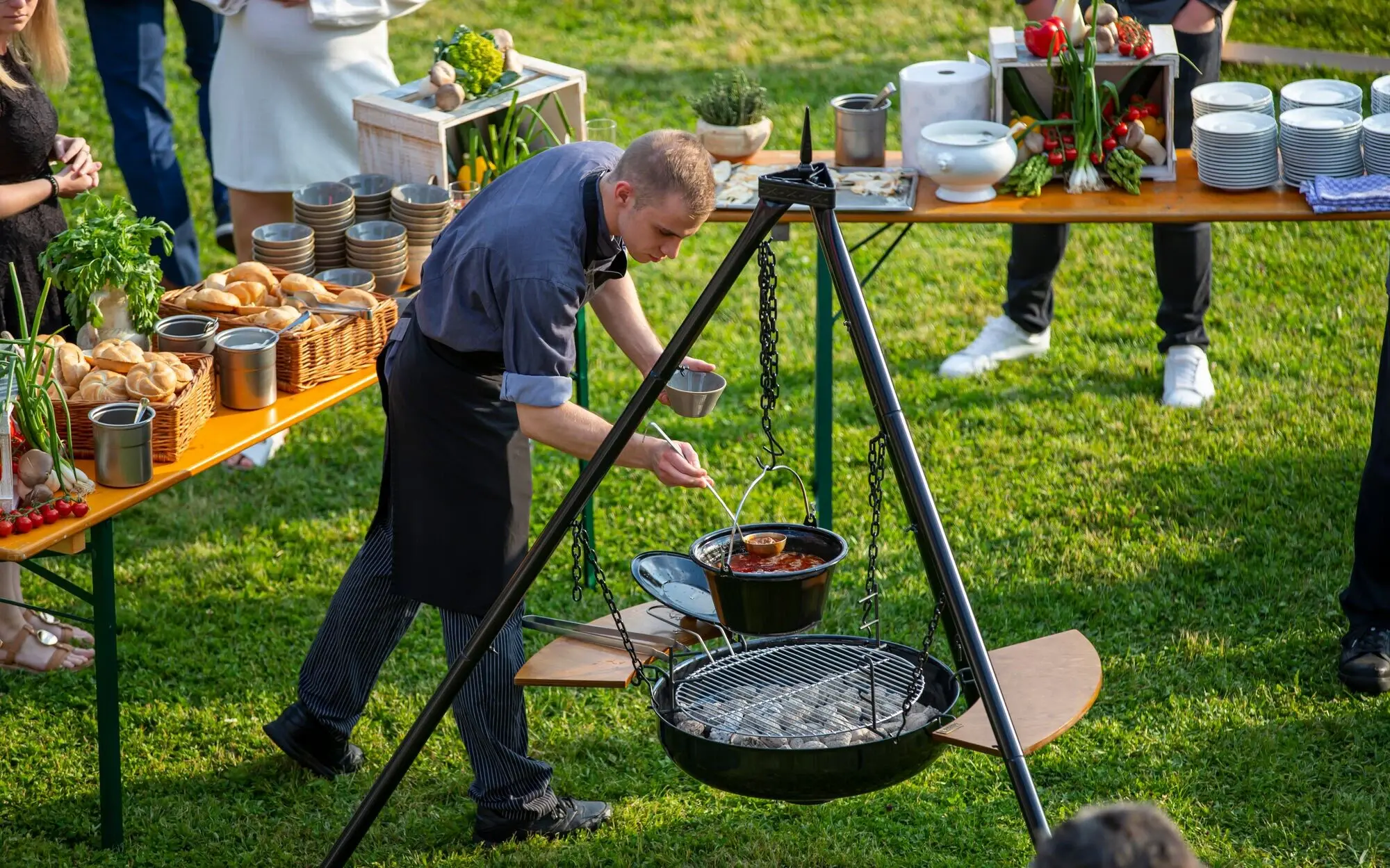 Koch steht an einem großem Dreibein Grill und kocht Essen im Freien.