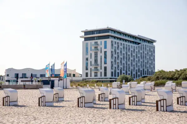 Eine Außenansicht des aja Warnemünde als weißes Gebäude mit vielen Fenstern an einem Strand mit weißen Strandkörben.