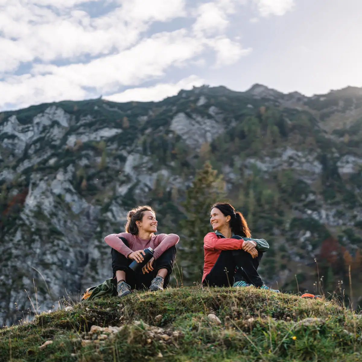 Wanderpause Zwei Frauen sitzen auf einem Hügel mit Bergen im Hintergrund.