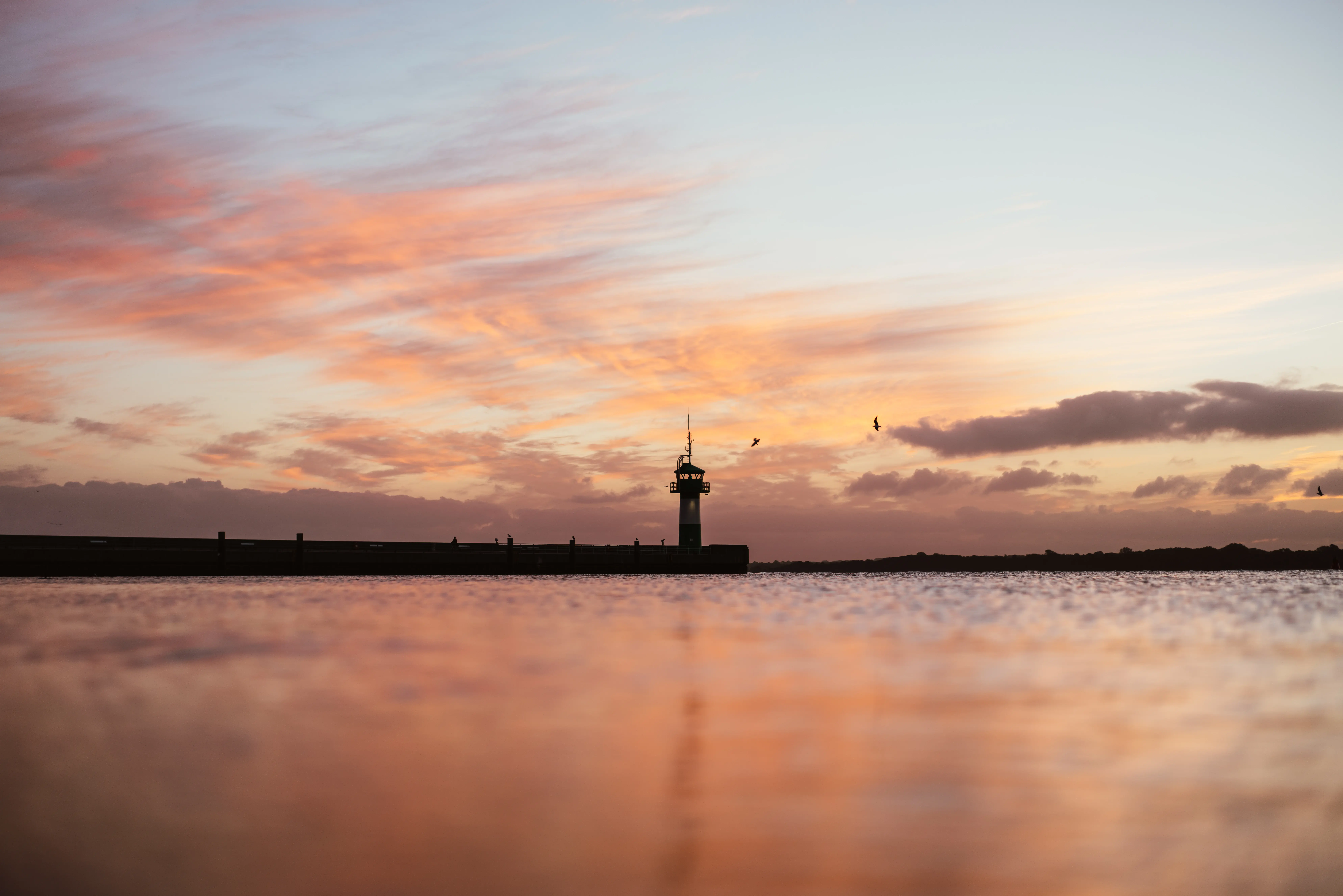 Ein langer Steg mit Leuchtturm am Ende führt auf ruhiges Wasser hinaus. Der leicht bewölkte Himmel ist in warme Farben der aufgehenden Sonne getaucht, die sich auch auf der Wasseroberfläche widerspiegeln.