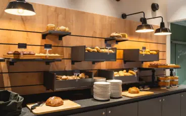 Eine warm beleuchtete Buffetstation mit Brot und Brötchen auf verschiedenen Regalebenen vor einer Holzwand.