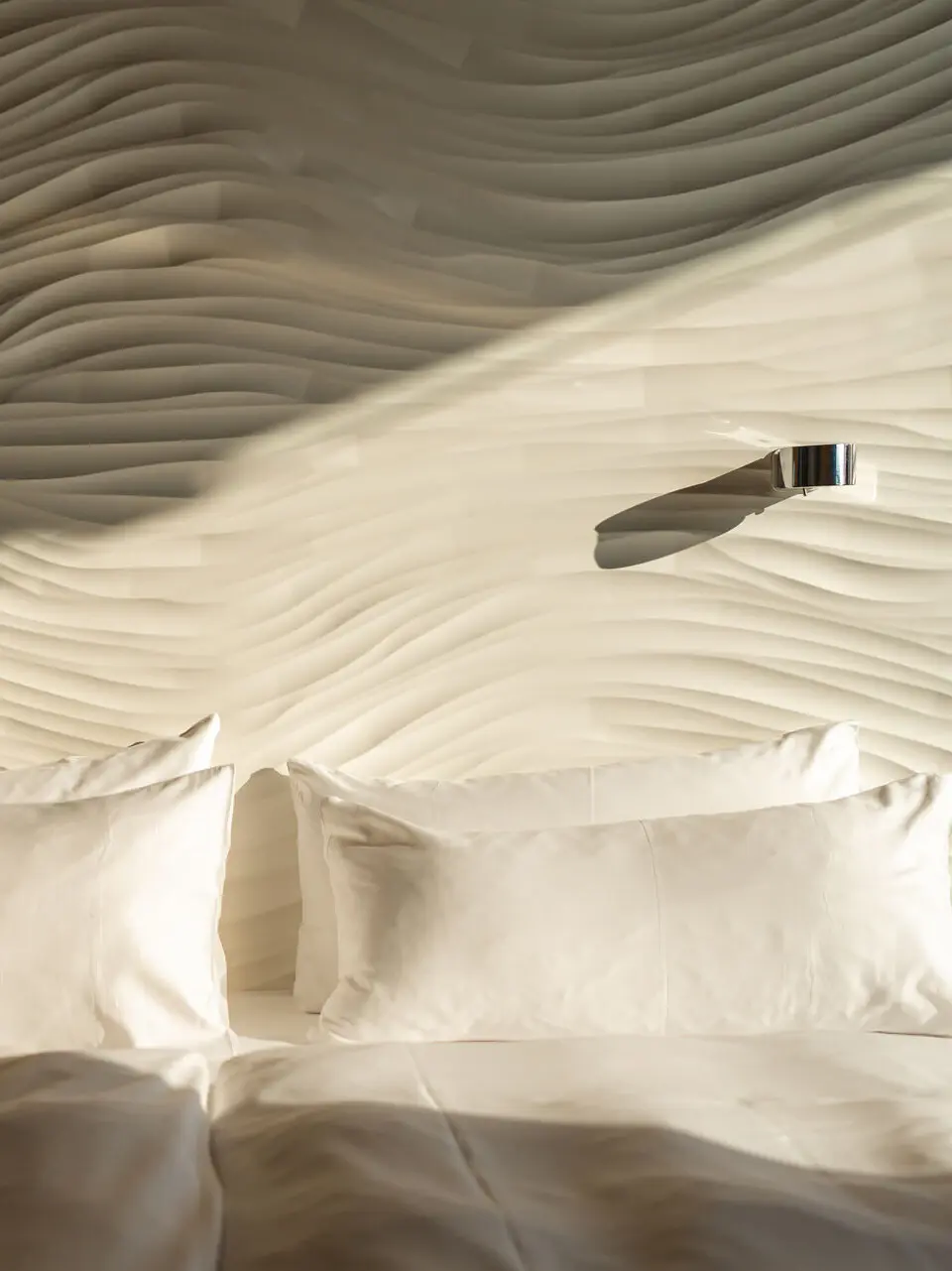 Eine Nahaufnahme von einem Bett und dem weiß welligen Kopfteil, an dem kleine runde Leselampen angebracht sind.