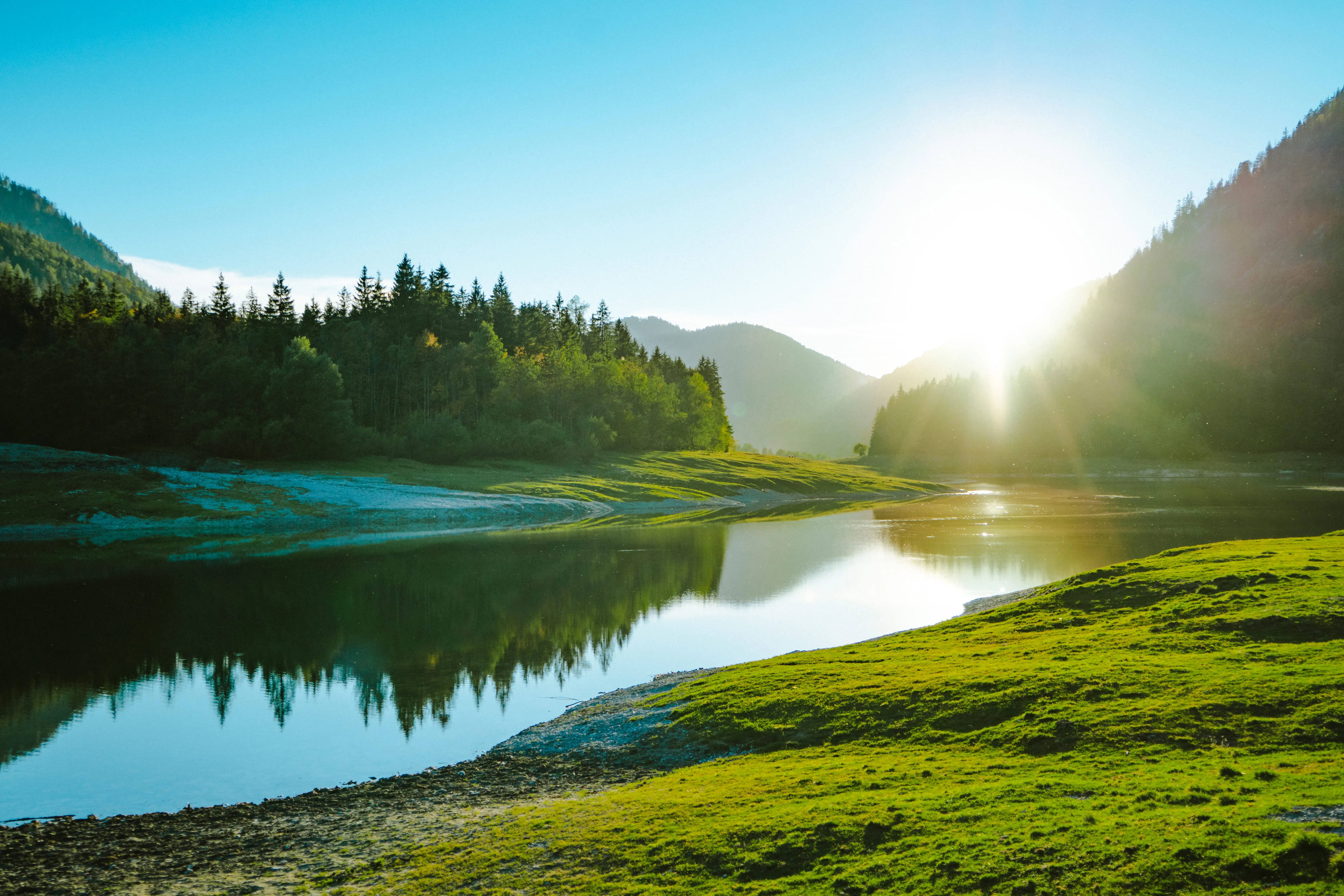 Destination_Ruhpolding Sonnenaufgang über einem ruhigen Bergsee, umgeben von grünen Wiesen und dichten Wäldern, mit Spiegelung der Landschaft im Wasser.