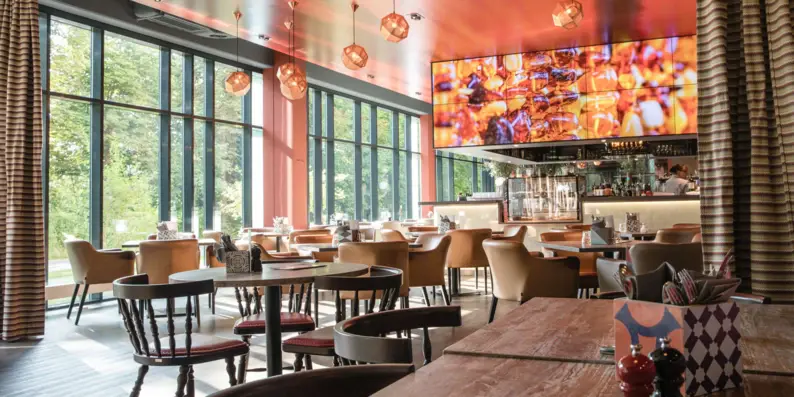 Innenraum eines Restaurants mit Tischen und Stühlen. Die große Fensterfront im Hintergrund füllt den Raum mit Tageslicht und über dem Tresen hängt eine große Anzeigetafel mit Essensabbildungen. Innenraum eines Restaurants mit Tischen und Stühlen. Die große Fensterfront im Hintergrund füllt den Raum mit Tageslicht und über dem Tresen hängt eine große Anzeigetafel mit Essensabbildungen.