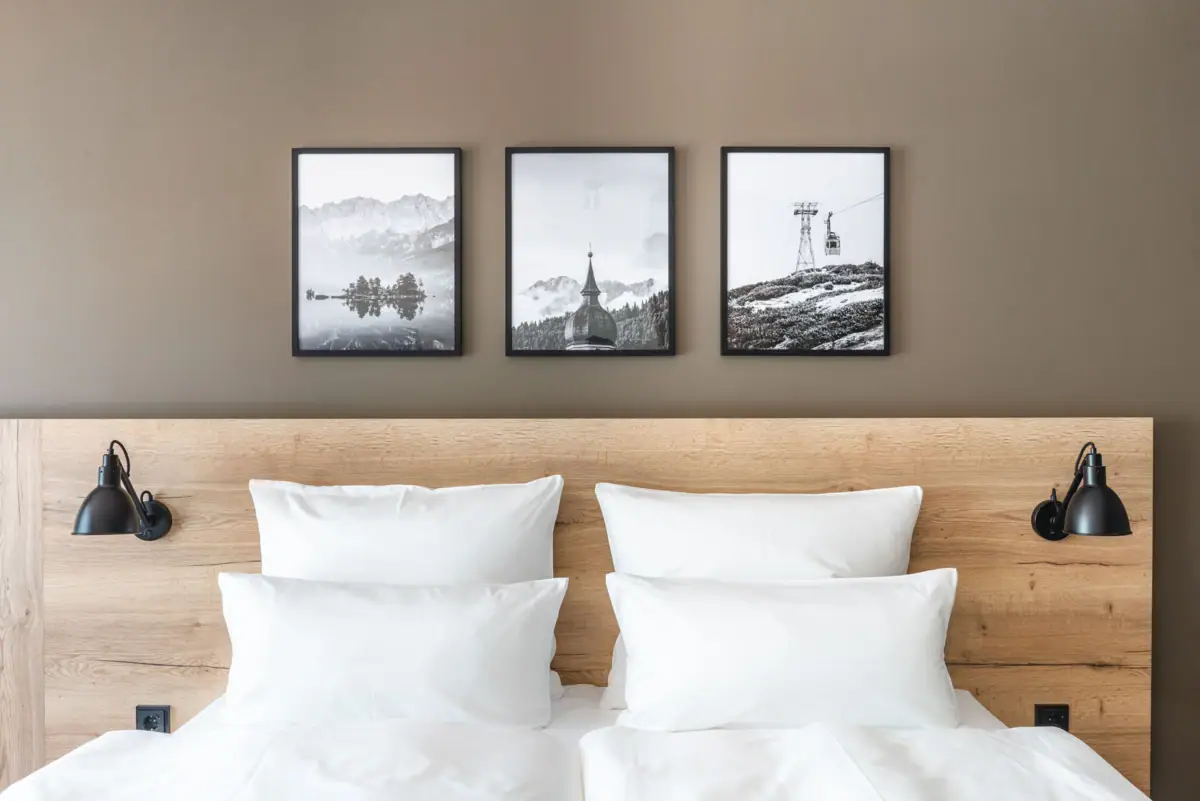 Zimmer Bett mit weißen Kissen und Holzrahmen, im Hintergrund ein Bild von einem Berg und einem Skilift.