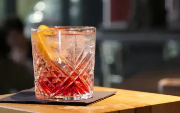 Ein Cocktail mit rotem in transparentem Farbübergang, Eiswürfeln und einer Orangenscheibe steht auf einer schwarzen Servierte auf einem Holztisch.