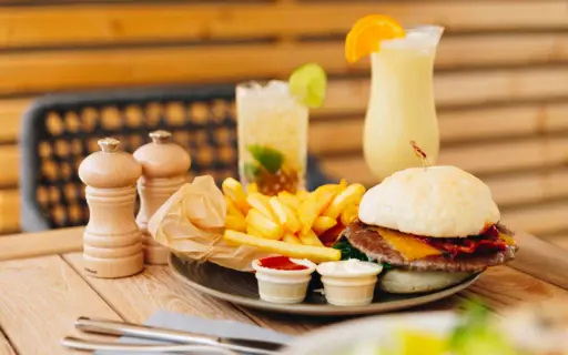 Leckeres Burger-Menü auf einem Holztisch, serviert mit Pommes und Dips. Im Hintergrund stehen zwei erfrischende Cocktails, dekoriert mit Limette und Orangenscheibe. Salz- und Pfefferstreuer ergänzen das Arrangement. Leckeres Burger-Menü auf einem Holztisch, serviert mit Pommes und Dips. Im Hintergrund stehen zwei erfrischende Cocktails, dekoriert mit Limette und Orangenscheibe. Salz- und Pfefferstreuer ergänzen das Arrangement.